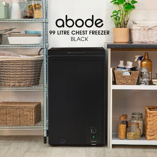 A black Abode 99 litre chest freezer stands in a kitchen-like setting between metal shelving and a white side table. The freezer has a small control panel and a green power light near the bottom right.