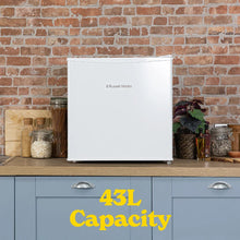 A white Russell Hobbs 43L mini refrigerator stands on a wooden countertop against a brick wall, with blue cabinets below and decorative jars and a plant on either side. Yellow text at the bottom reads '43L Capacity'.