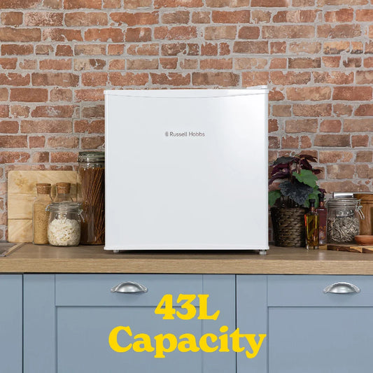 A white Russell Hobbs 43L mini refrigerator stands on a wooden countertop against a brick wall, with blue cabinets below and decorative jars and a plant on either side. Yellow text at the bottom reads '43L Capacity'.